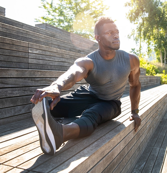 A man stretches his arms and legs while seated on a wooden bench in a park setting. A man stretches his arms and legs while seated on a wooden bench in a park setting.
