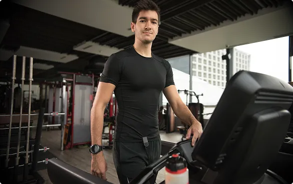 A man rides an exercise bike in a gym, surrounded by fitness equipment and bright overhead lights.