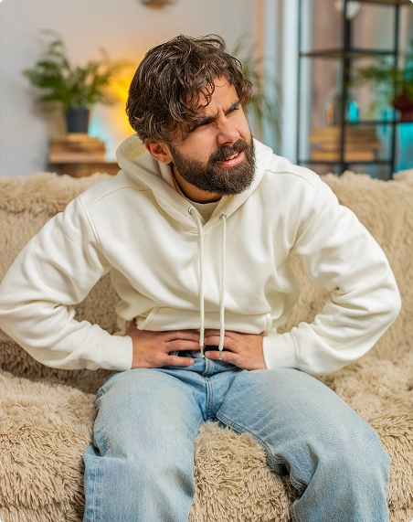 A man sitting on a couch, grimacing in discomfort, holds his stomach as he experiences pain A man sitting on a couch, grimacing in discomfort, holds his stomach as he experiences pain