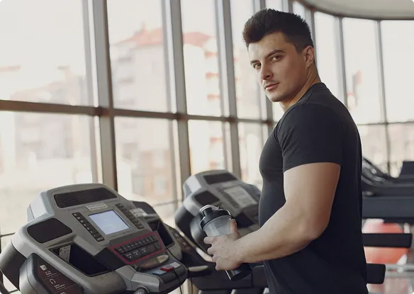 A man is using a treadmill in a gym, concentrating on his fitness routine with other gym-goers in the background.