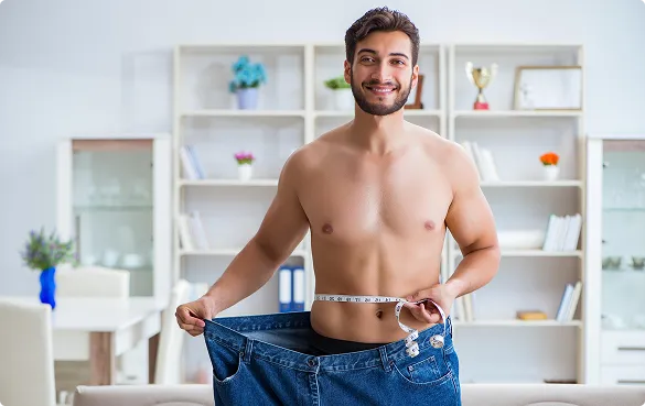 A shirtless man displays his jeans and uses a measuring tape to check the fit around his waist.