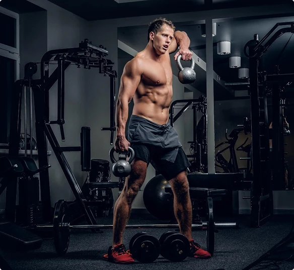 A muscular, shirtless man holds a kettlebell in a gym, demonstrating his fitness routine and dedication.