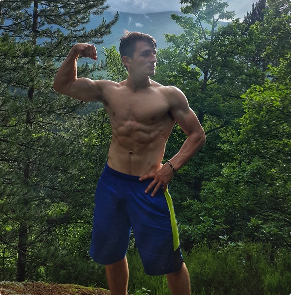 A young man poses with one arm flexed, showcasing his muscular physique against a backdrop of trees and mountains. The scene is set in a natural, outdoor environment.