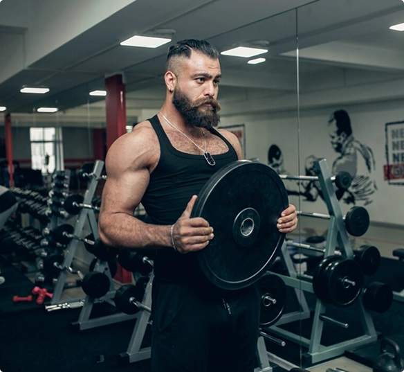 A bearded man holds a plate while standing in a gym, surrounded by fitness equipment.