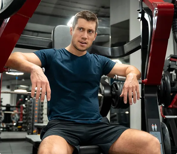 A man seated on a gym bench, surrounded by exercise equipment in a fitness center.