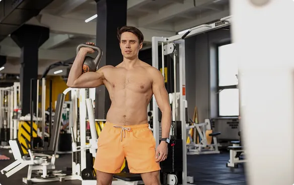  A shirtless man wearing an orange swimsuit holds a dumbbell, emphasizing his fitness and strength in a gym setting.