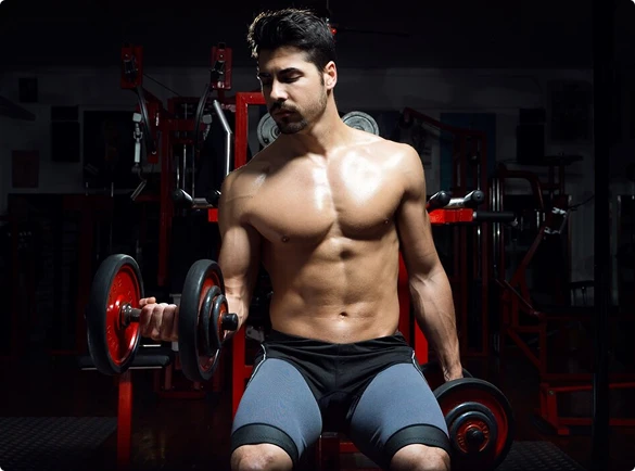 A shirtless man relaxes on a bench in a gym, surrounded by weights and fitness equipment, indicating a workout session.