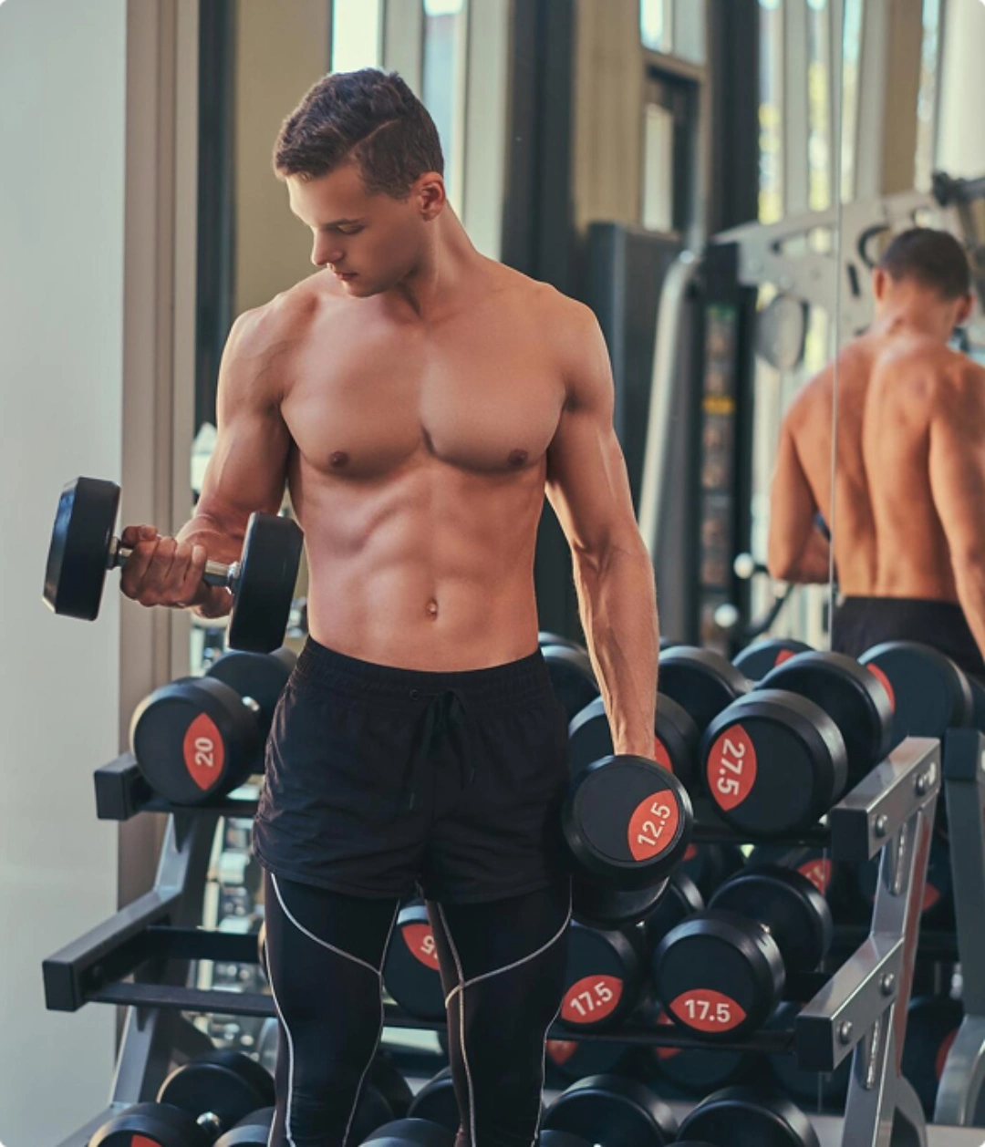 A shirtless man lifts two dumbbells while exercising in a gym setting.