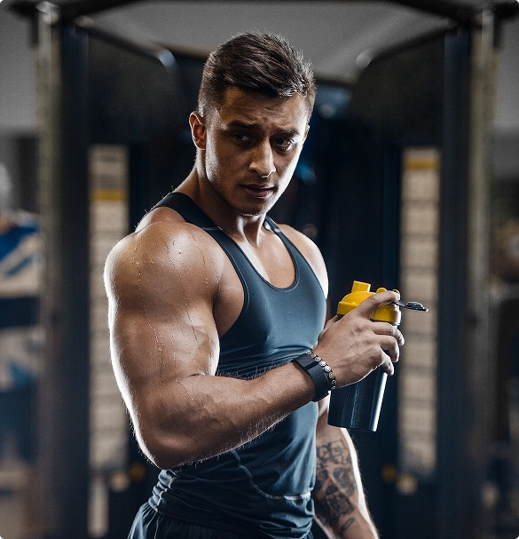 A muscular man in a black tank top holds a shaker bottle and sports drink, standing in a gym with fitness equipment in the background.