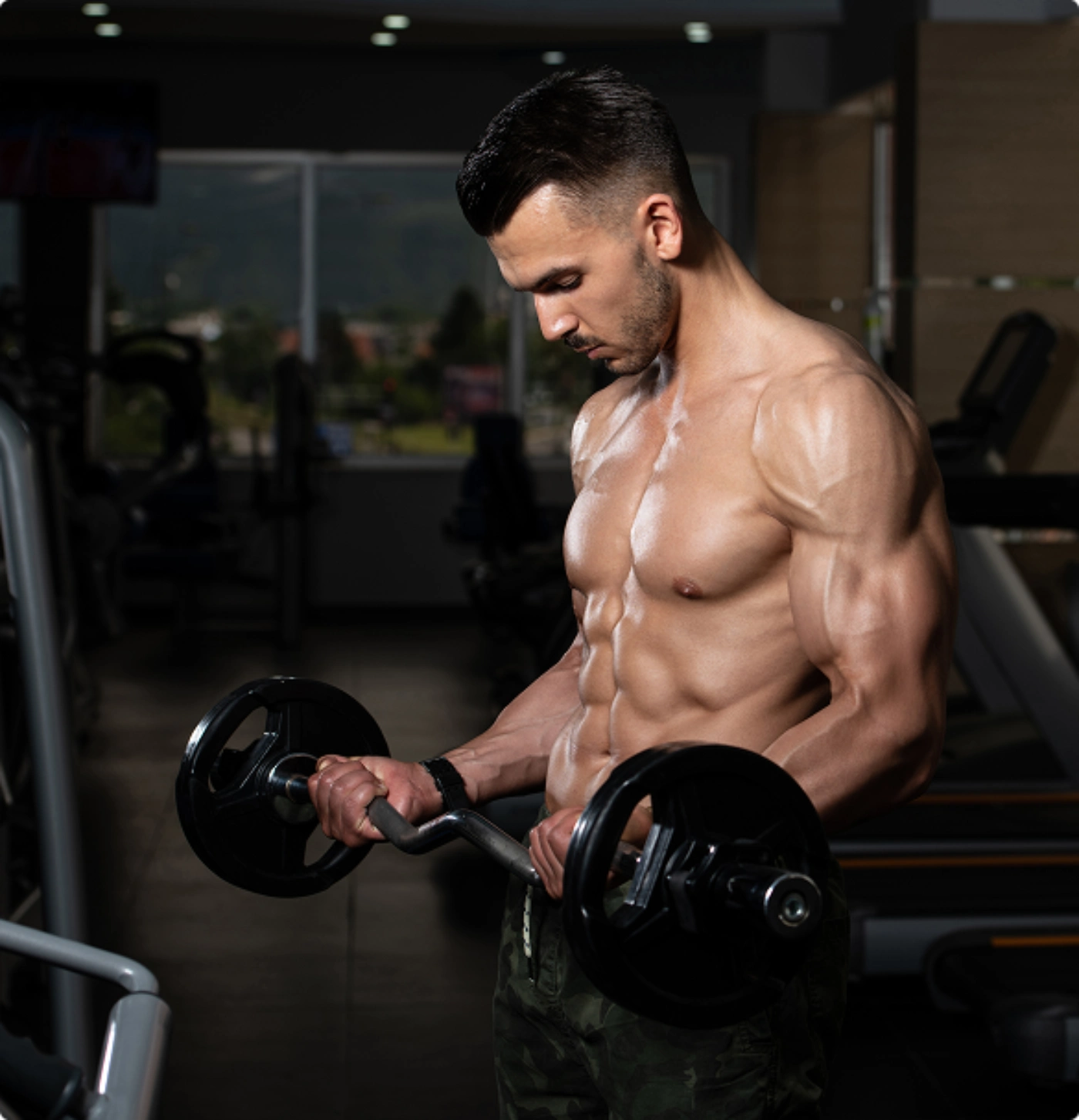 A muscular person lifts a barbell in a gym, showcasing defined abs and arms, with exercise equipment and a window in the background.
