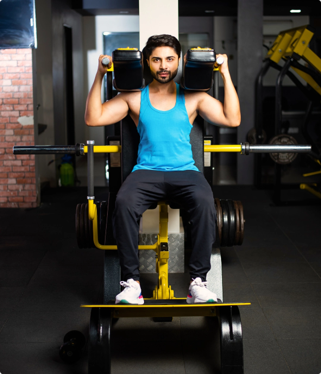 A person in a blue tank top performs a shoulder press on a gym machine, surrounded by weights in a modern fitness center.