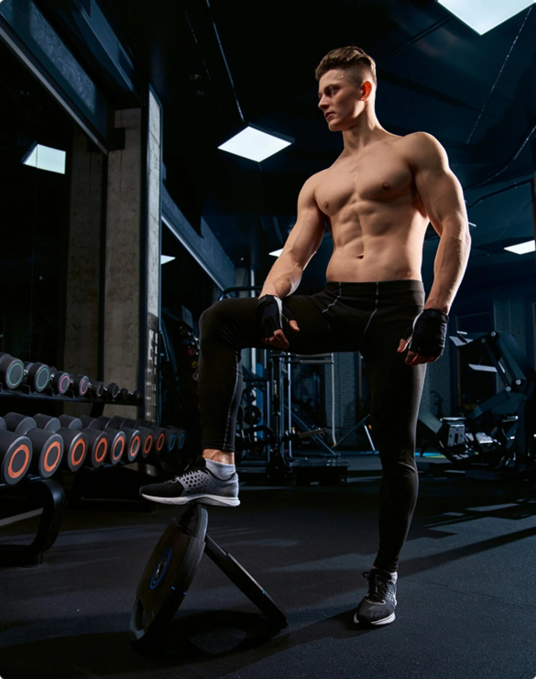 A muscular man stands confidently on a weight plate in a modern gym, showcasing strength and dedication in a well-lit environment.