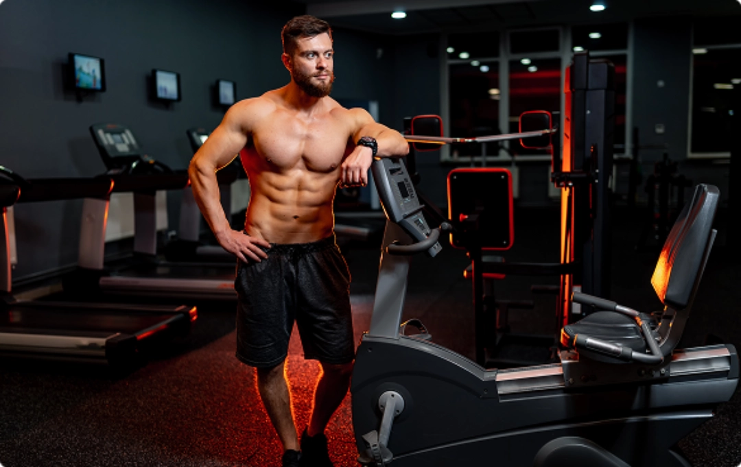 A muscular person leans against gym equipment in a well-lit fitness center, showcasing an active lifestyle and workout environment.