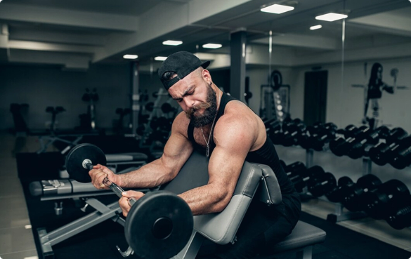 A man wearing a cap and tank top performs bicep curls with a barbell on a preacher bench in a gym with various weights and equipment in the background.