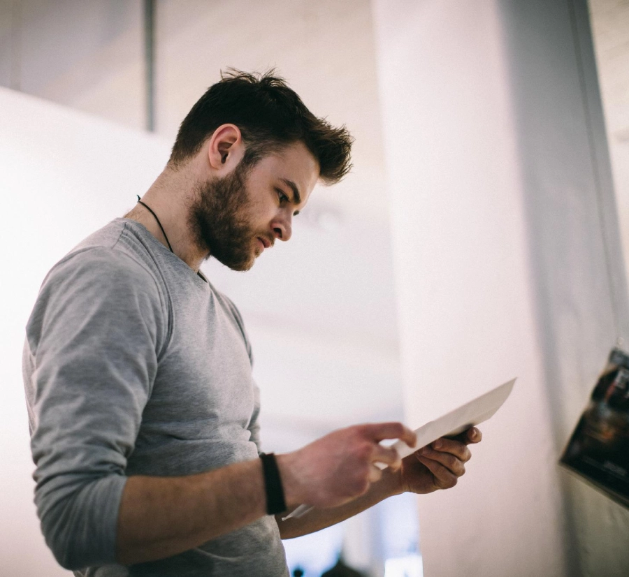 A man stands before a white wall, intently examining a piece of paper in his hands.